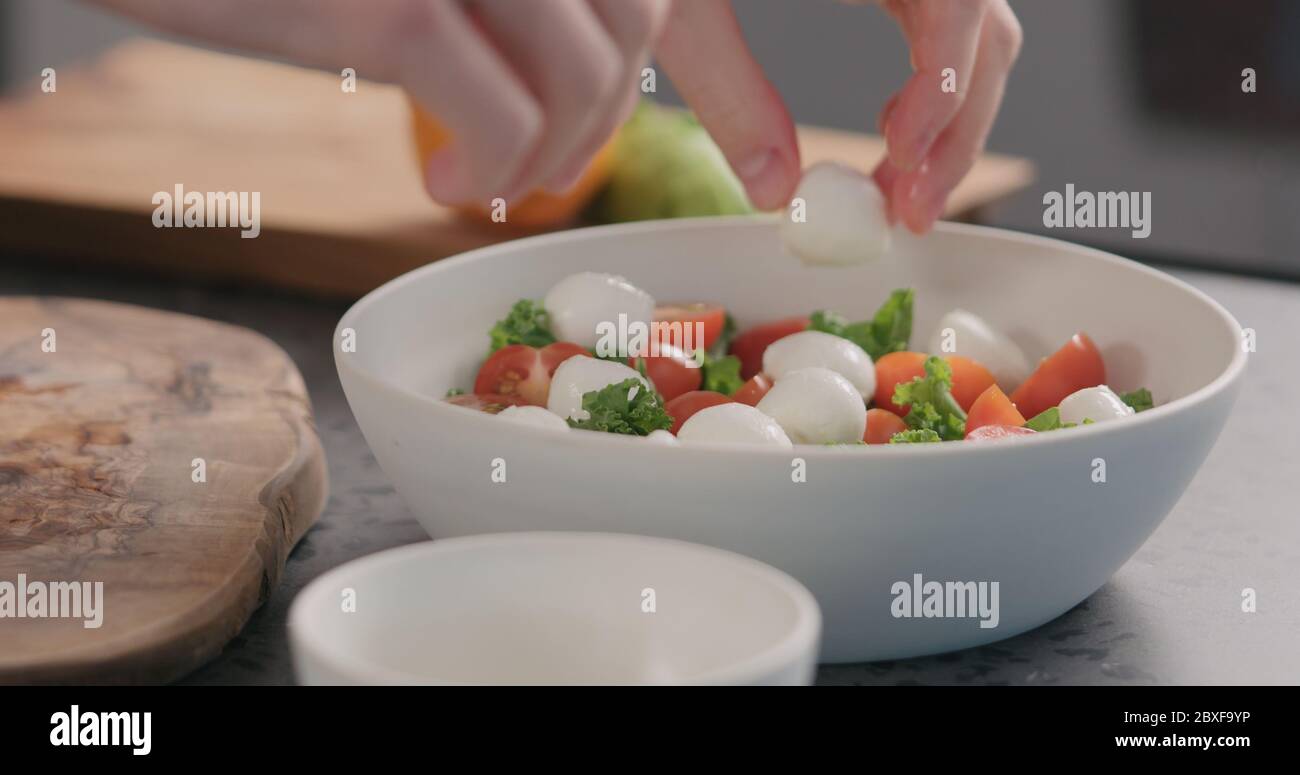 l'uomo aggiunge mozzarella nell'insalata con kale e pomodori ciotola bianca sul piano cottura Foto Stock