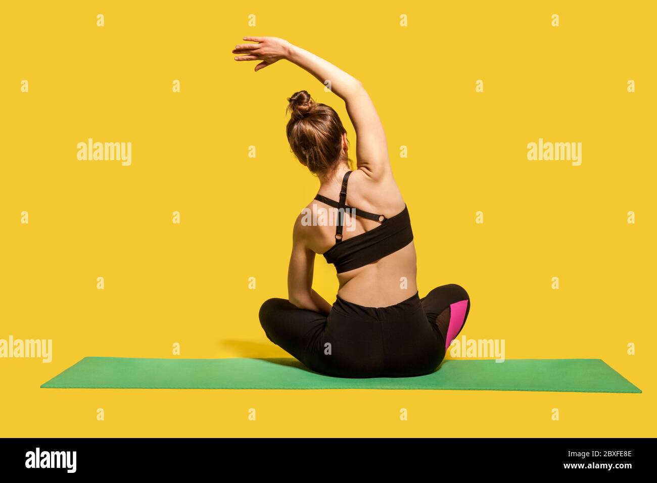 Vista posteriore atleta donna con capelli in tight top e pantaloni seduta in posizione lotus sul tappetino palestra praticare yoga, piegandosi a fianco con la mano sollevata, d Foto Stock