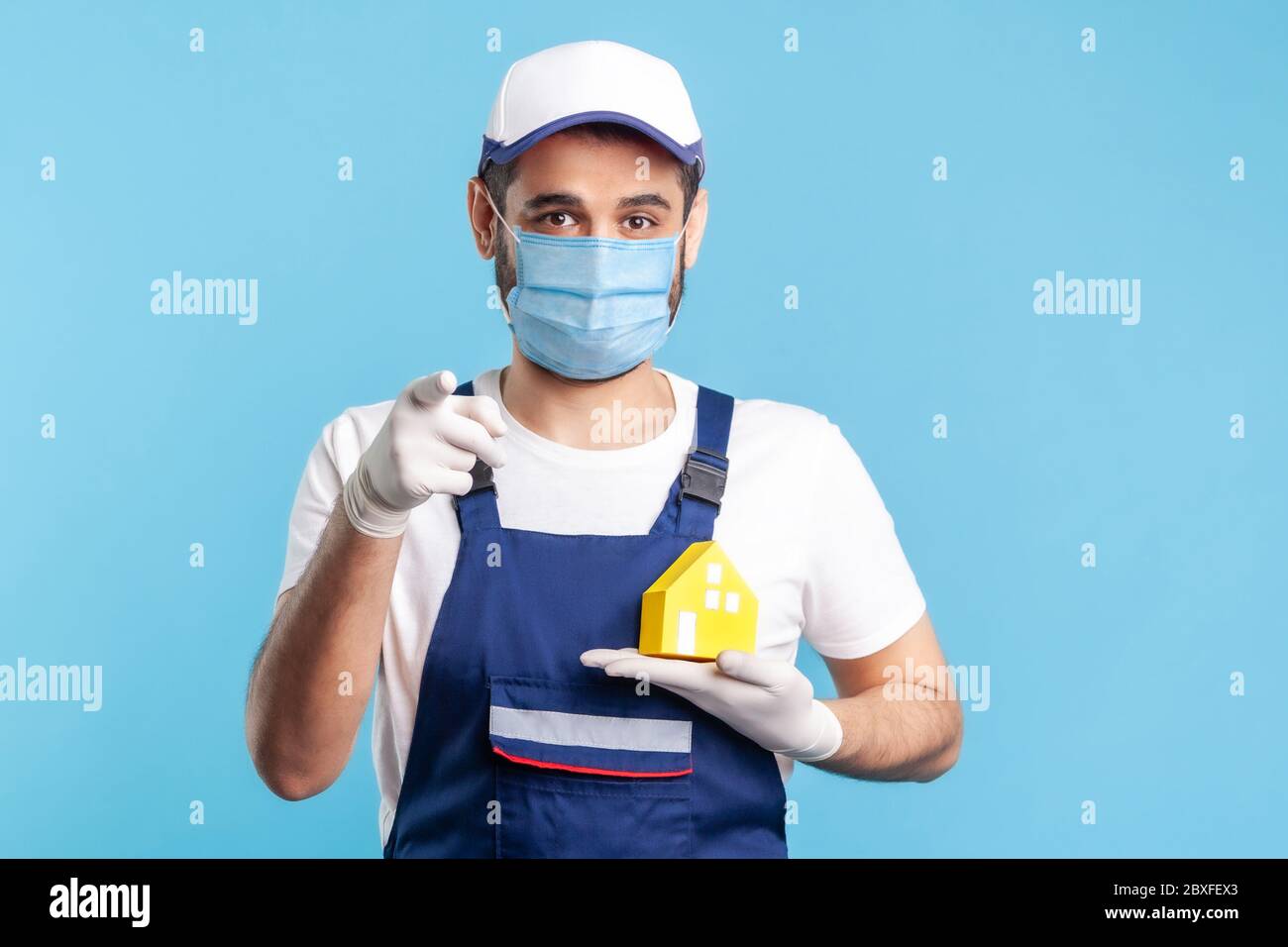 Ehi te! Ritratto di felice handyman, uomo in movimento in abbigliamento da lavoro, maschera e guanti che tengono la casa di carta e che punta alla macchina fotografica. Pubblicità riparazione casa, maint Foto Stock