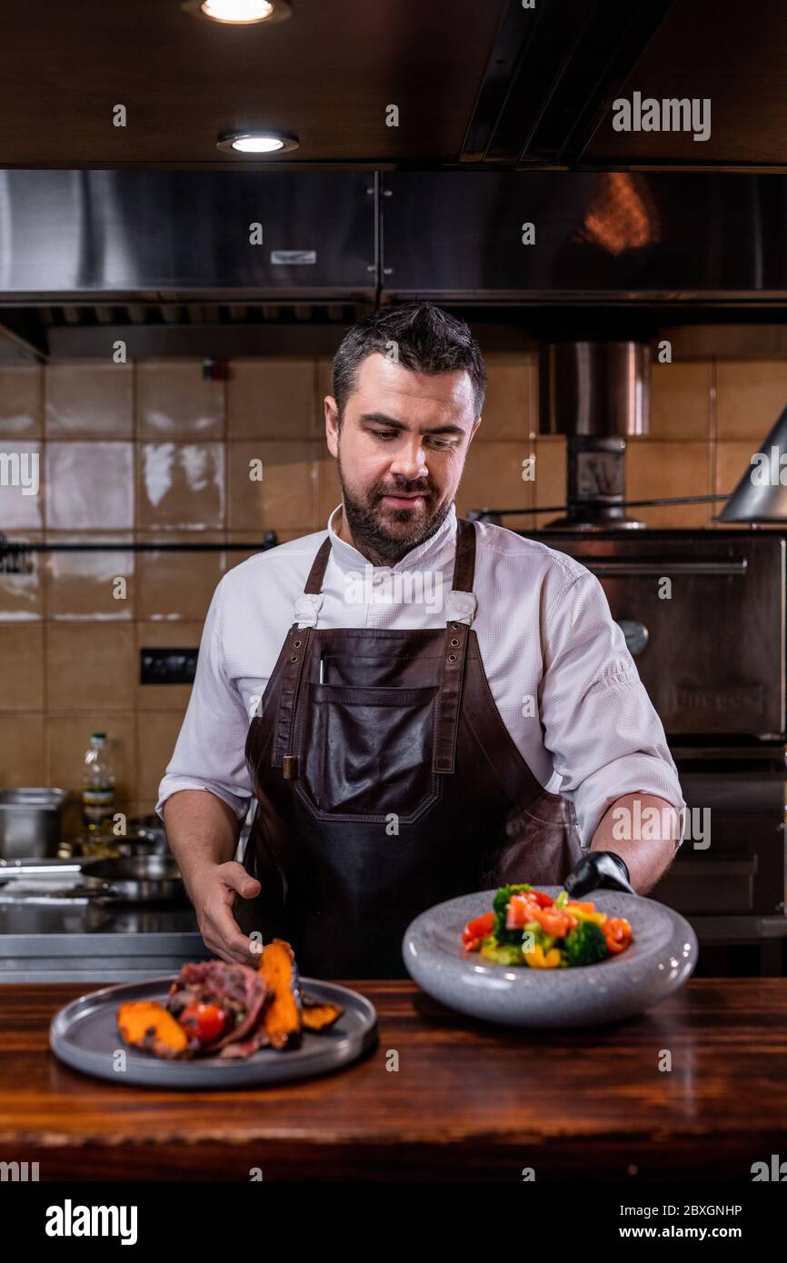Bel cuoco in grembiule di pelle in piedi al banco e che serve piatti nel ristorante Foto Stock