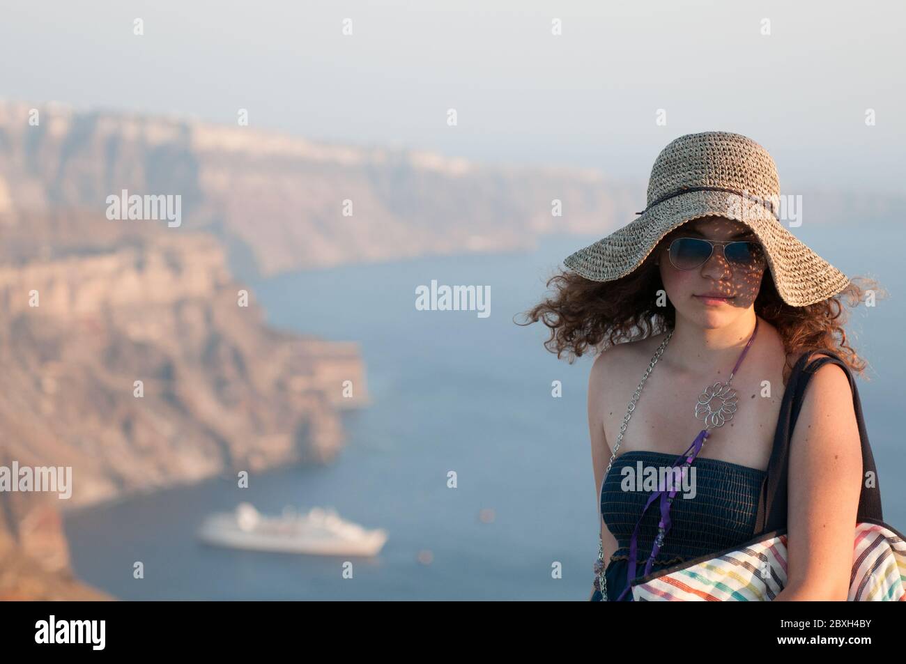 Bella ragazza felice che indossa un cappello con vista caldera dietro a Santorini Island in Grecia. Vacanze estive nelle isole greche Foto Stock