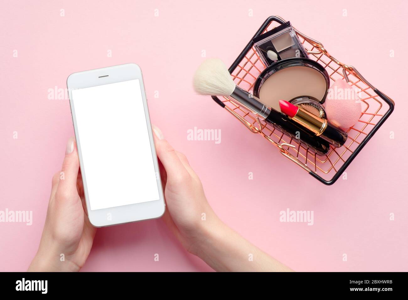 Acquisto di prodotti di bellezza online concetto. Donna mano che tiene telefono cellulare con schermo vuoto mockup e carrello con cosmetici su sfondo rosa t Foto Stock