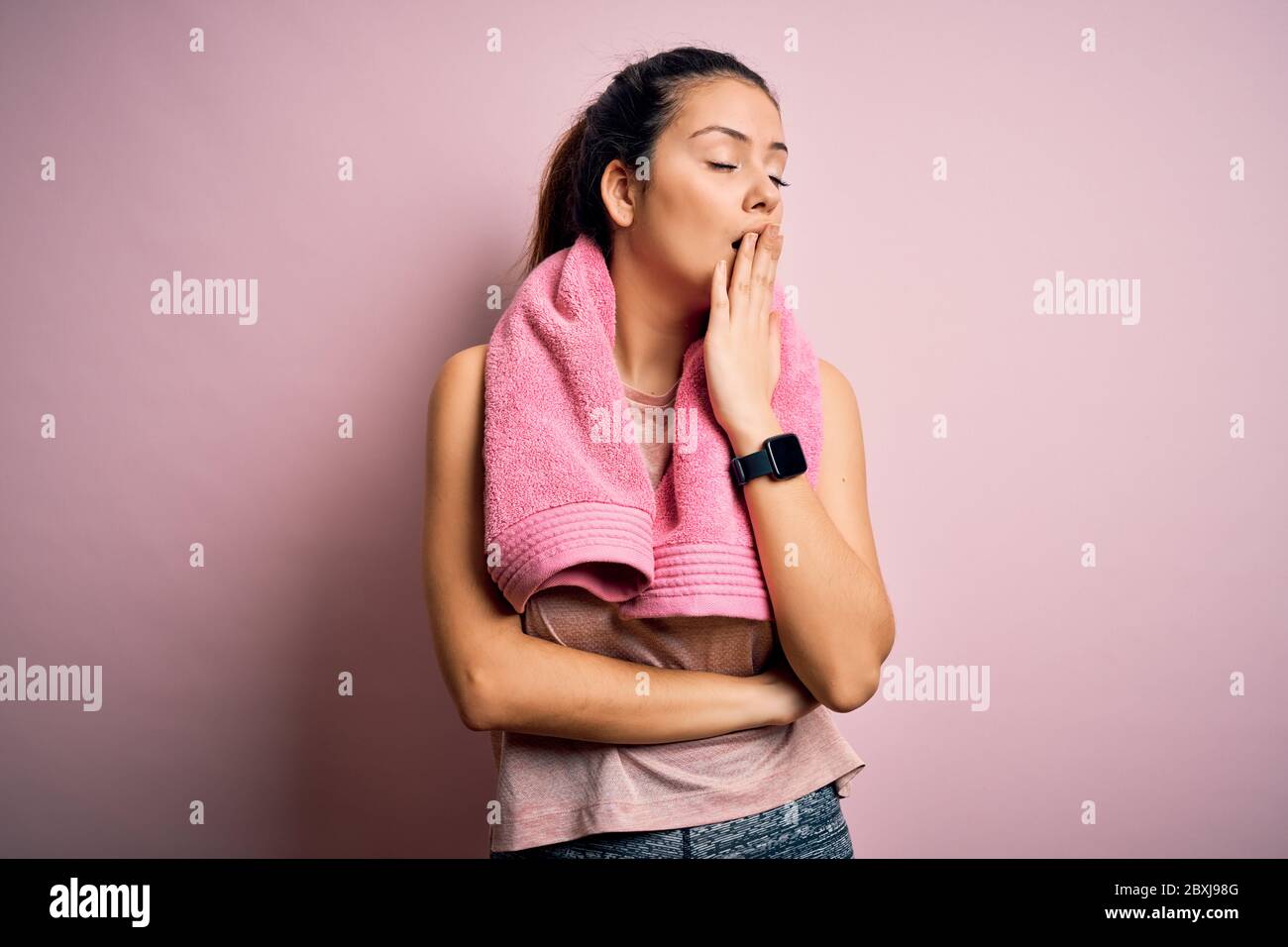 Giovane bella bruna sportivo donna con indosso sportswear e asciugamano su sfondo rosa annoiato tendente stanco coprendo bocca con mano. Irrequieto e sled Foto Stock