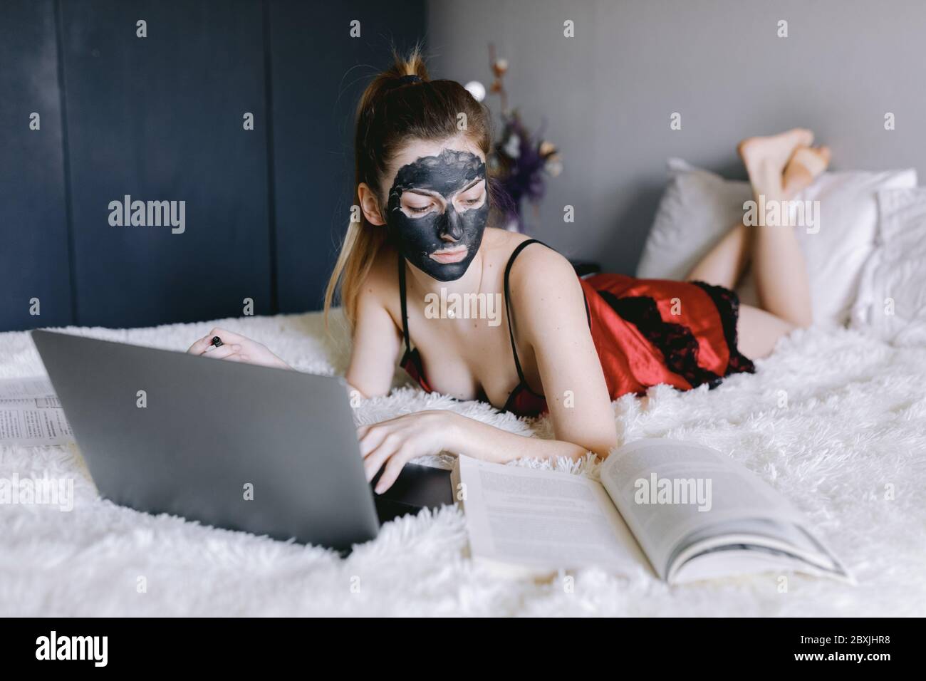 Studente con maschera di fango che legge il libro di testo e utilizza il laptop Foto Stock