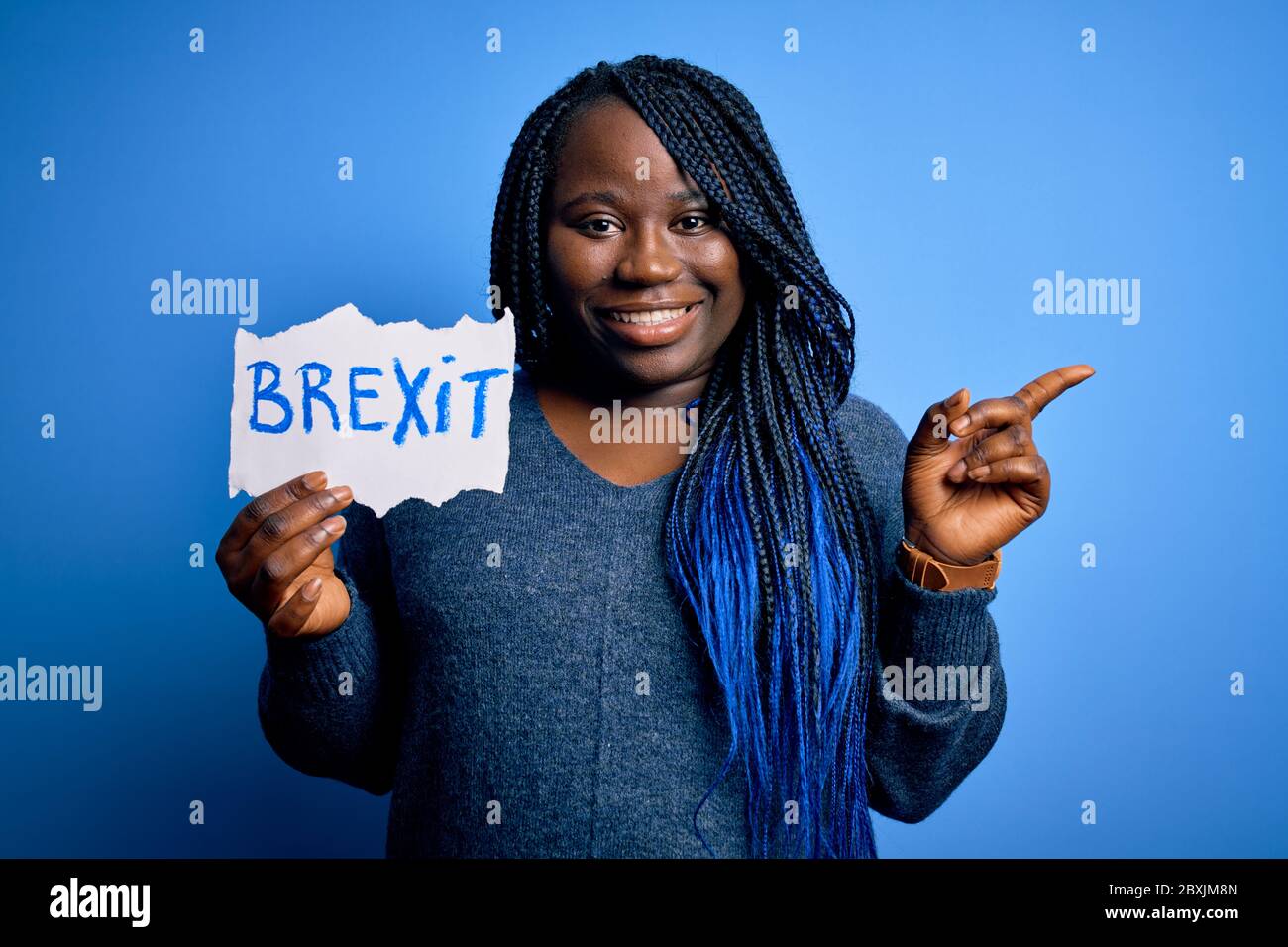 Giovane afroamericano più taglia donna con trecce tenendo carta con brexit messaggio molto felice puntando con mano e dito a lato Foto Stock