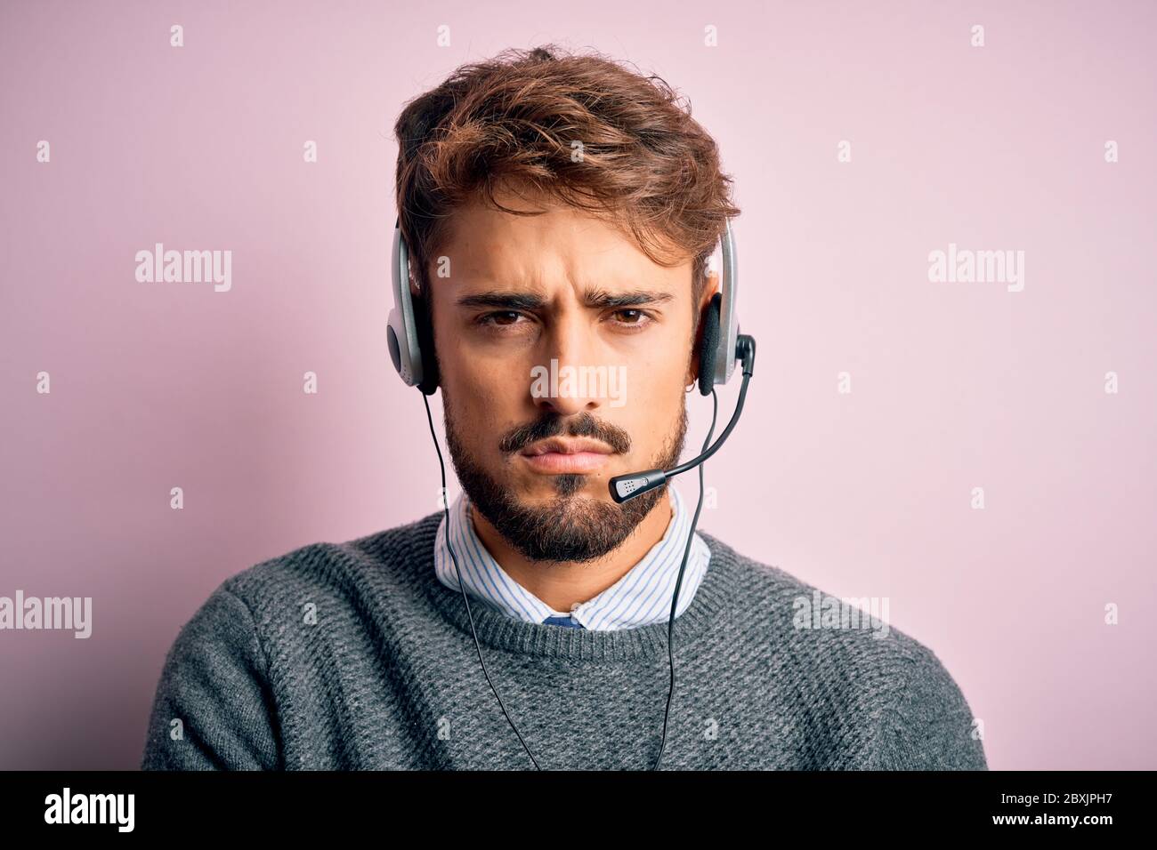 Giovane agente del call center uomo con barba che indossa cuffie su uno sfondo rosa isolato, scettico e nervoso, disapprovando l'espressione sul viso con croce Foto Stock