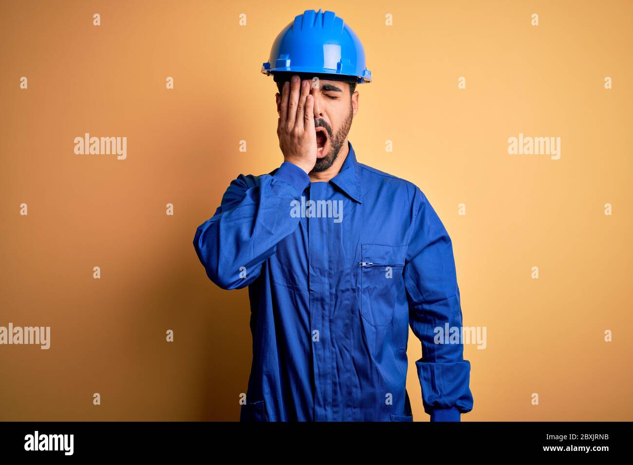 Uomo meccanico con barba che indossa uniforme blu e casco di sicurezza su sfondo giallo che si stanca coprendo metà faccia, occhio e bocca con la mano. Faccia Foto Stock