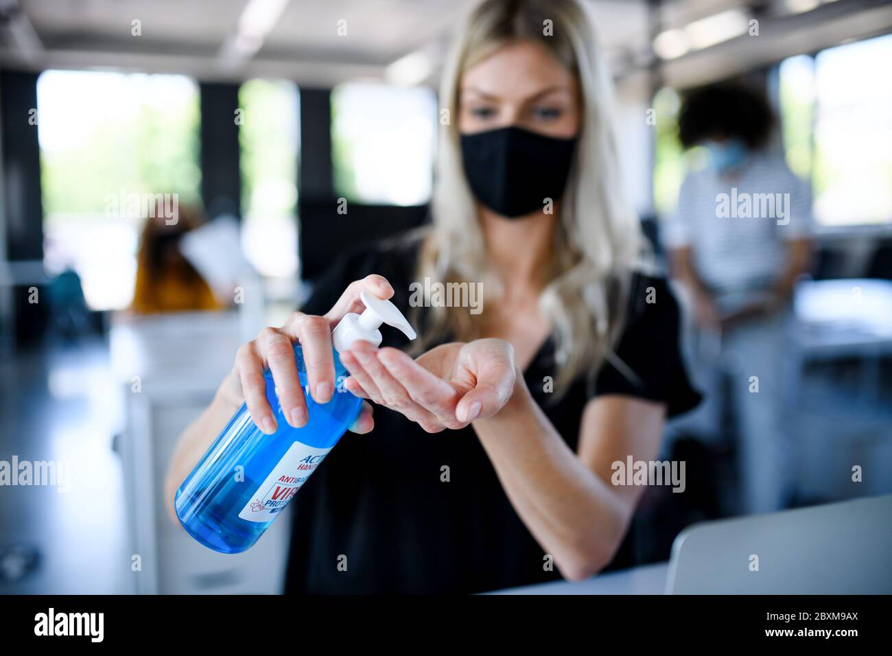 Giovane donna con maschera facciale sul lavoro in ufficio dopo il blocco, disinfettando le mani. Foto Stock