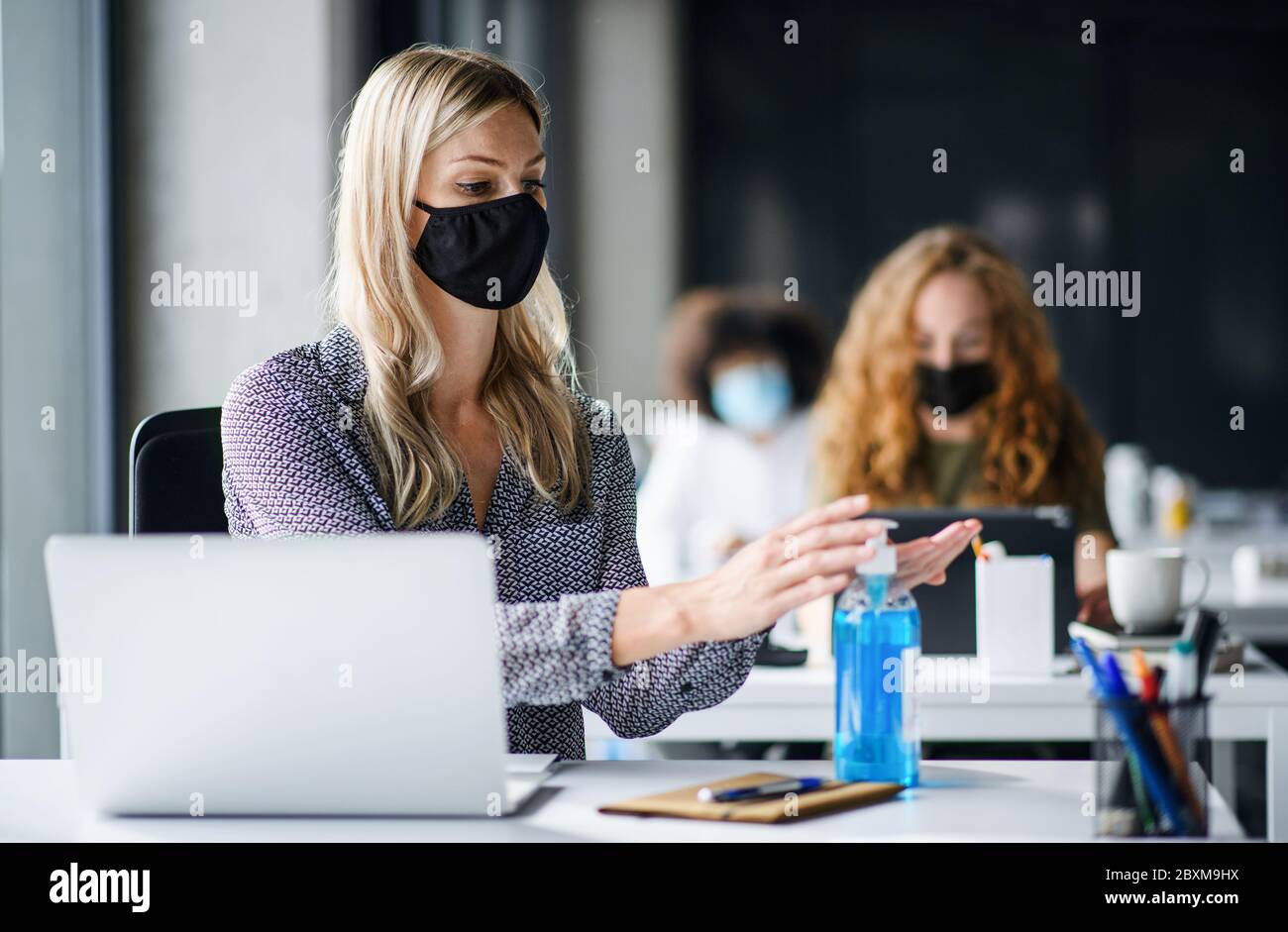 Giovane donna con maschera facciale sul lavoro in ufficio dopo il blocco, disinfettando le mani. Foto Stock