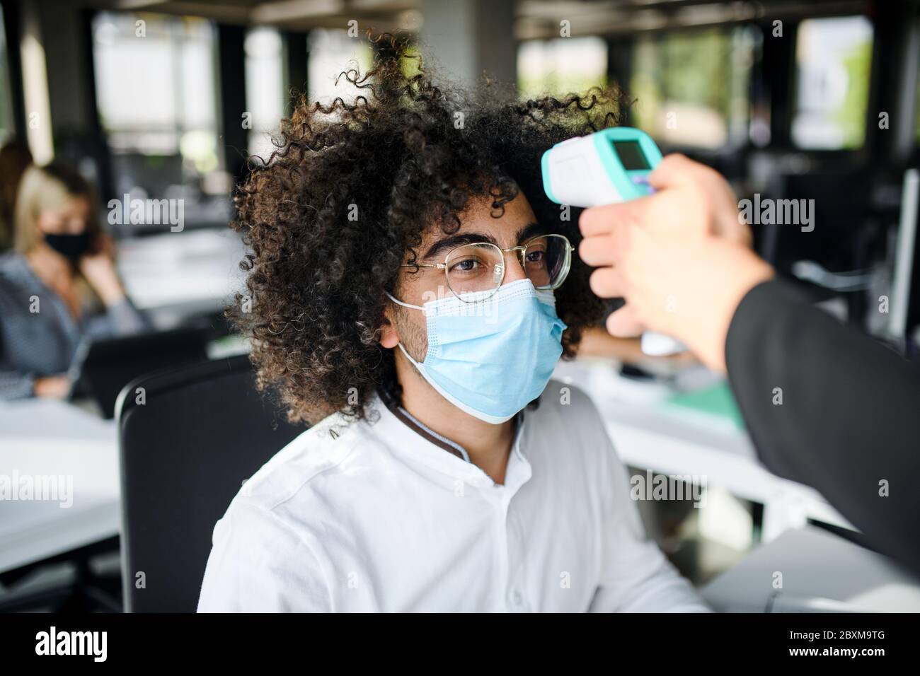 Giovane uomo con maschera facciale torna al lavoro in ufficio dopo il blocco, misurando la temperatura. Foto Stock