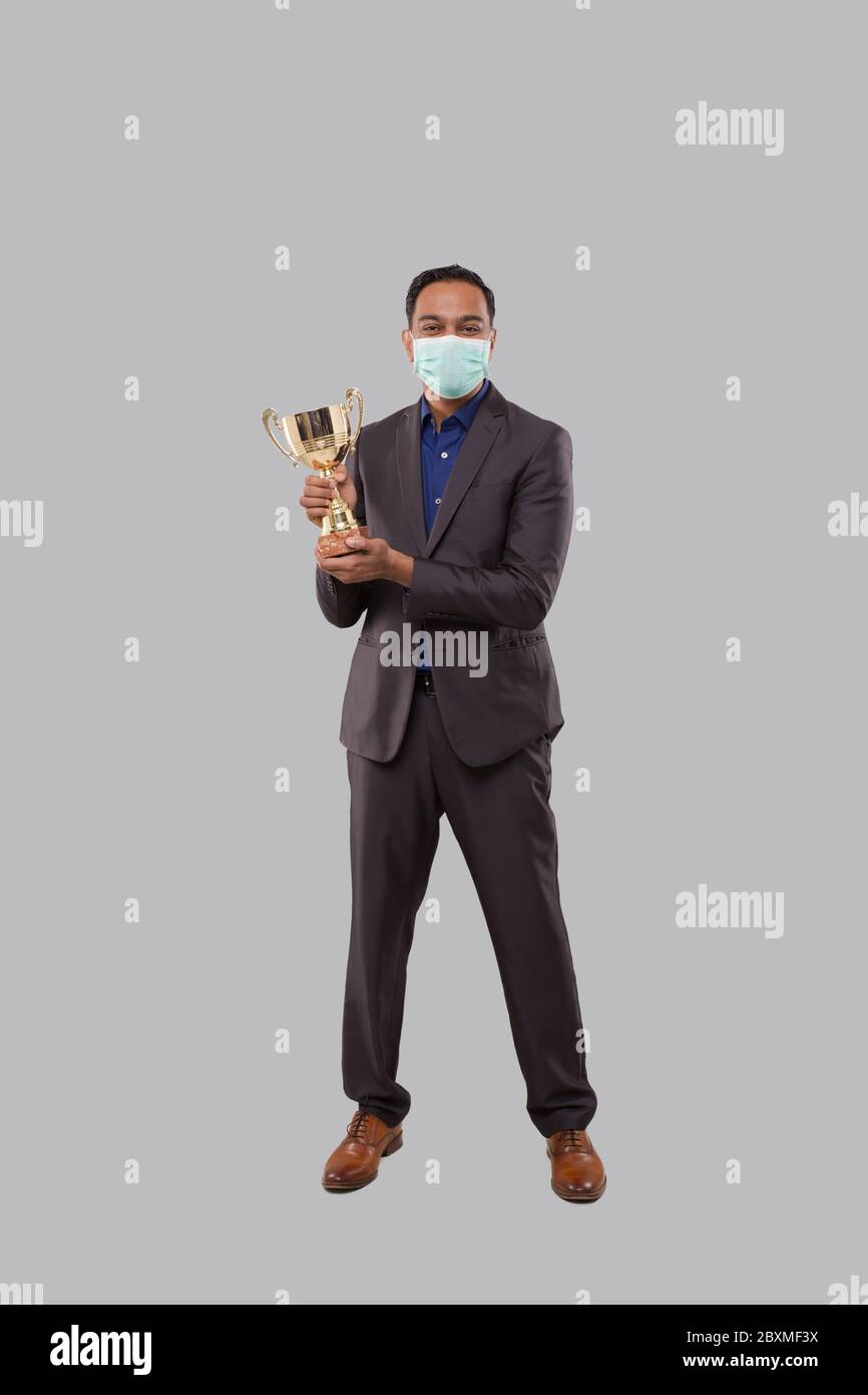 Businessman Holding Trophy indossare maschera medica. Uomo d'affari indiano in piedi a lunghezza intera con Trofeo in mani Foto Stock