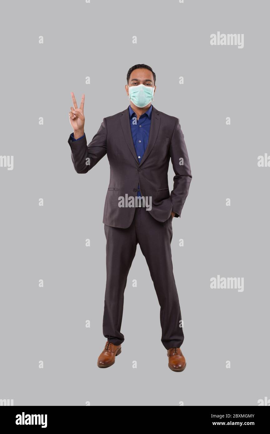 Uomo d'affari che mostra un cartello di Pace che indossa una maschera medica. Indio uomo d'affari in piedi di lunghezza completa. Foto Stock