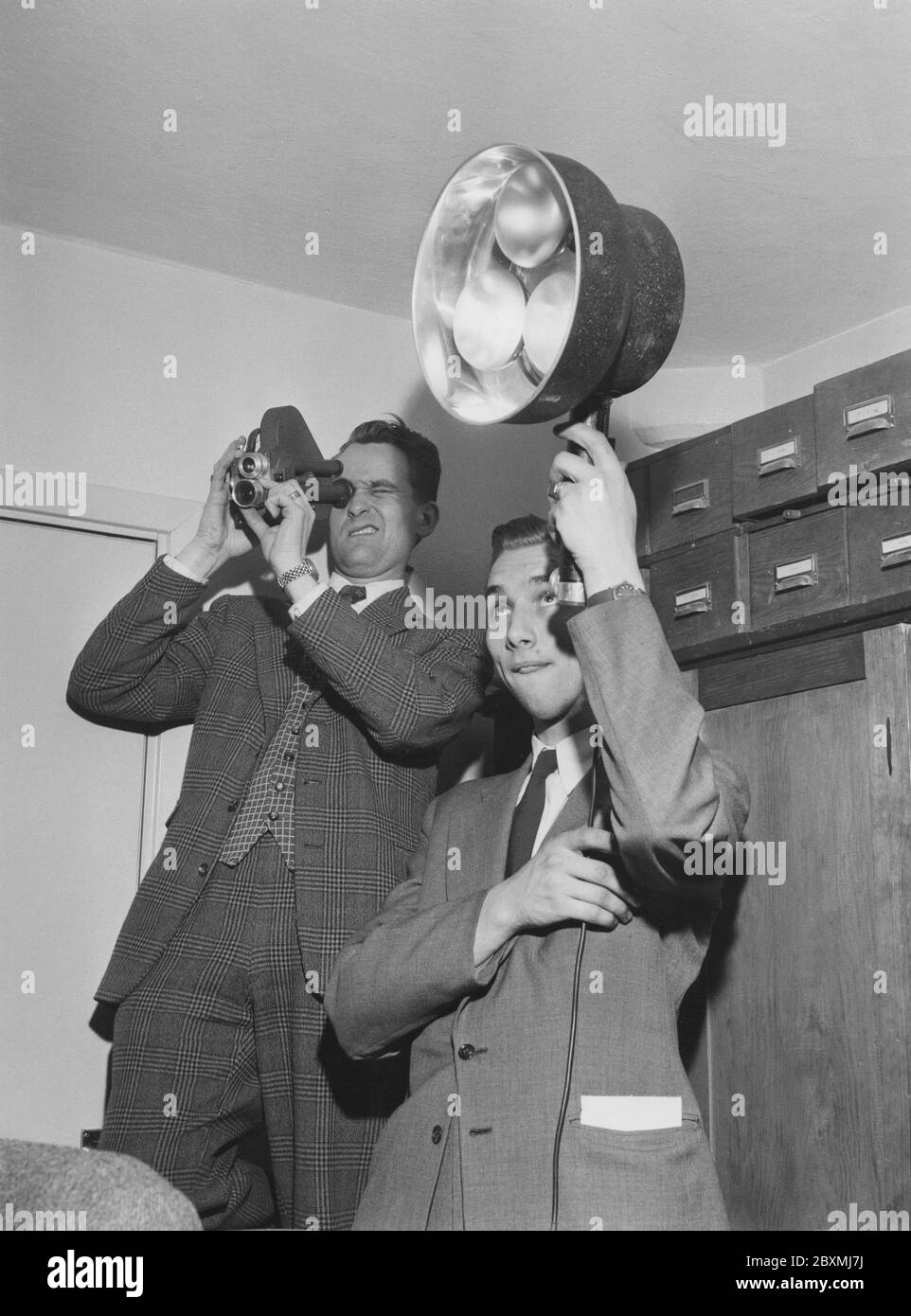 Riprese negli anni '50. Un uomo sta girando con una piccola macchina fotografica da 8 mm mentre il suo amico sta tenendo le lampade per illuminare il soggetto che viene girato. La telecamera ha prodotto una pellicola analogica che è stata visualizzata su uno schermo bianco da un proiettore. Svezia 1958 Foto Stock Riprese negli anni '50. Un uomo sta girando con una piccola macchina fotografica da 8 mm mentre il suo amico sta tenendo le lampade per illuminare il soggetto che viene girato. La telecamera ha prodotto una pellicola analogica che è stata visualizzata su uno schermo bianco da un proiettore. Svezia 1958 Foto Stock