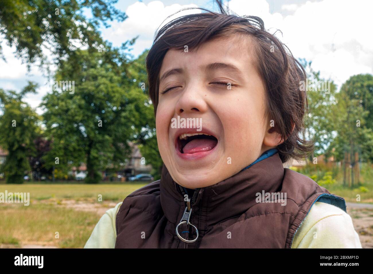 Un ritratto di un ragazzo con gli occhi chiusi e la bocca aperta catturati in mezzo alla risata, portato fuori nel parco locale Foto Stock