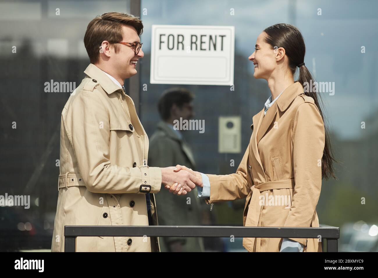 Vista laterale vita in su ritratto di sorridente agente immobiliare scuotendo le mani con il cliente all'aperto mentre si sta accanto a PER il CARTELLO DI AFFITTO Foto Stock