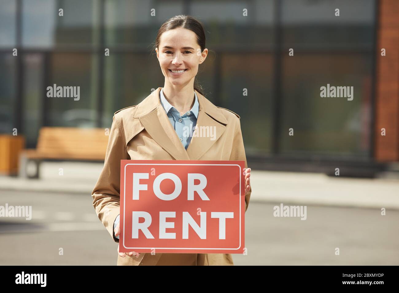 Vita in su ritratto di agente immobiliare femminile sorridente guardando felice la macchina fotografica mentre tiene rosso PER L'AFFITTO segno all'aperto, spazio di copia Foto Stock