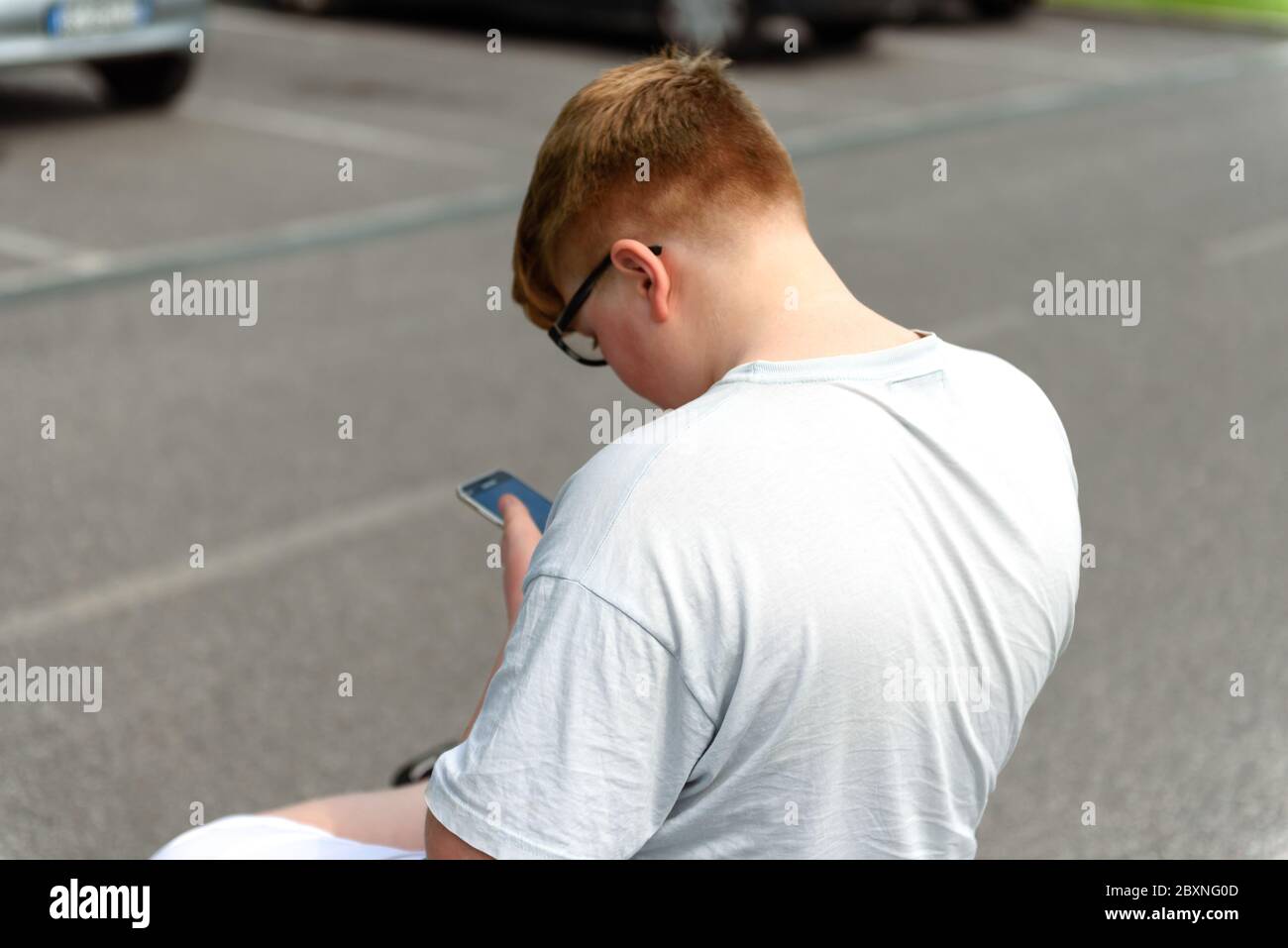 Vista da dietro di un ragazzo dai capelli rossi con cellulare e occhiali in un contesto urbano. Gli occhiali da ragazzo consentono di tenere lo smartphone a portata di mano. Foto Stock