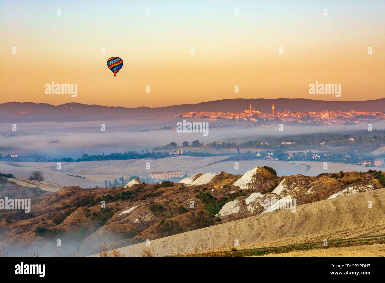 Un colorato pallone sospeso nell'aria che galleggia nel cielo di prima mattina tra il paesaggio delle crete senesi e la città di Siena sullo sfondo Foto Stock