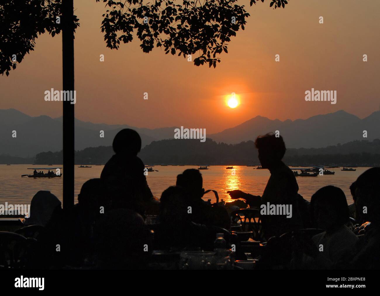 Il tramonto sul Lago Ovest (Xi Hu) a Hangzhou, nella provincia di Zhejiang, Cina. Vista al tramonto sul Lago Ovest, Hangzhou, con le sagome di persone sedute in un cafe. Foto Stock