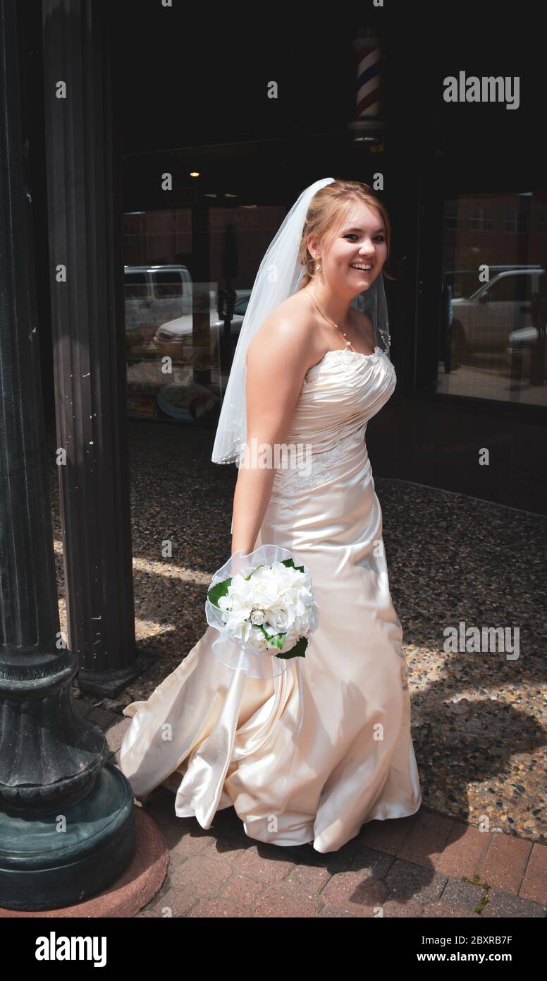 Una giovane sposa caucasica in abito da sposa bianco senza maniche con velo, sorride tenendo il bouquet mentre si cammina da solo lungo un marciapiede nel centro di St. Cloud, Minnesota Foto Stock