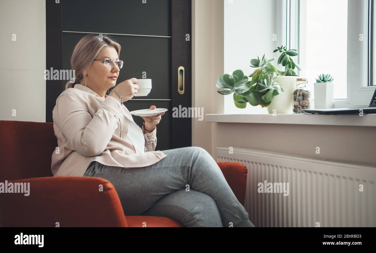 Donna caucasica anziana con occhiali seduti in poltrona e bere un caffè mentre si indossa occhiali e guardare una lezione sul computer portatile Foto Stock