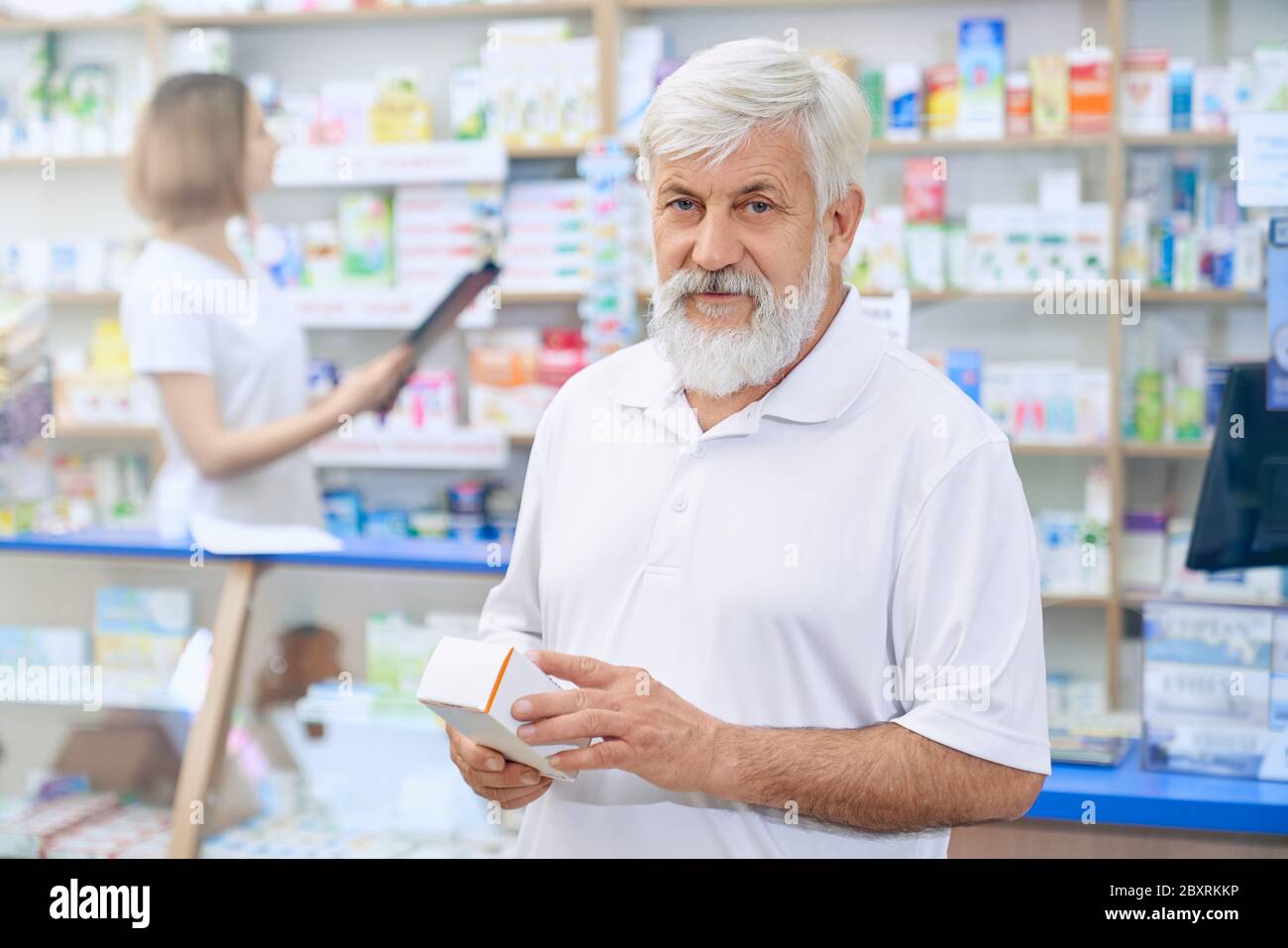 Ritratto di uomo anziano che sceglie farmaci in farmacia, guardando la macchina fotografica. Cliente senior con bearded in camicia bianca con scatola medica. Farmacista femminile in piedi al banco sullo sfondo. Foto Stock