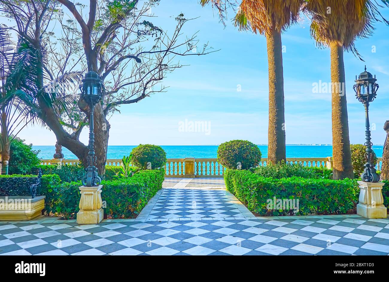 Il giardino perfettamente paesaggistico fronte oceano del Paseo de Carlos III con cespugli topiari, alte palme, retrò luci della strada in ghisa e scacchiera di pietra pa Foto Stock