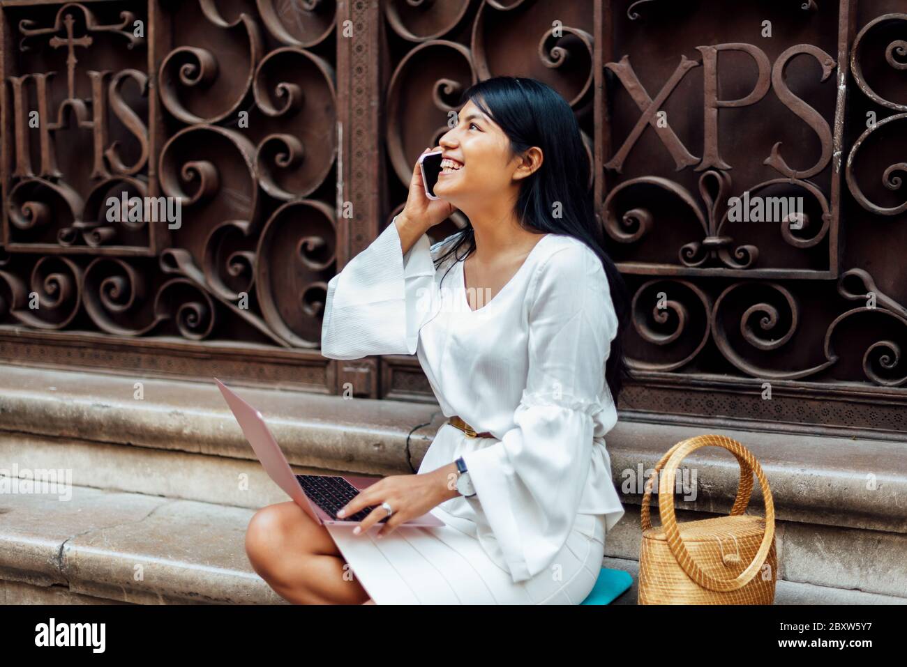 Donna latina brunette con lunghi capelli seduti su scala in pietra con computer portatile e parlando al telefono mentre sorride. Concetto donna digitale Foto Stock