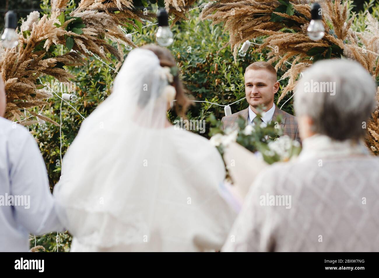Sposo in piedi vicino all'arco di nozze e guardando la sposa che lo conduce. Sposa e sposo. Cerimonia nuziale Foto Stock