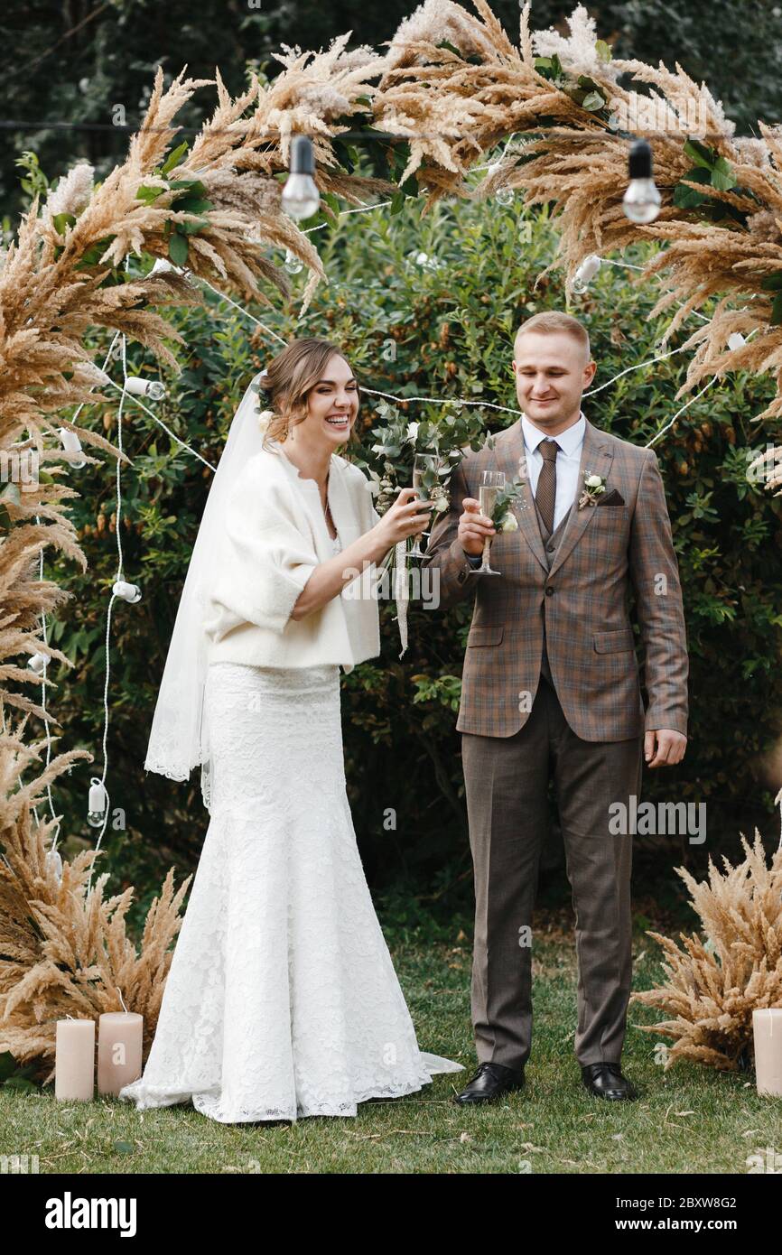 Sposa sorridente e sposo in piedi vicino all'arco del matrimonio con il bicchiere di champagne in mano. Sposa e sposo Foto Stock