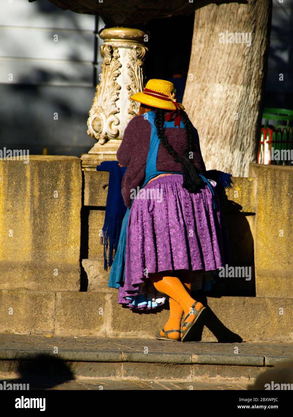 Donna boliviana - colita - in tipico abito colorato con gonna viola e capanna gialla in piedi alla fontana. Vista dal lato posteriore. Potosi, Bolivia Foto Stock