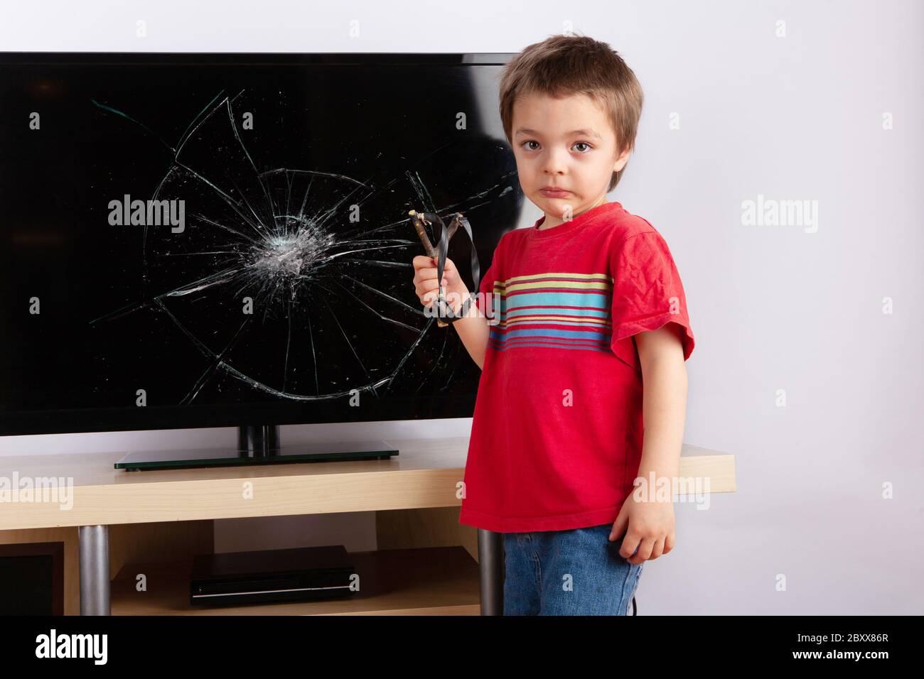 Un ragazzino spaventoso che si trova di fronte a una TV con schermo rotto che tiene una foto. Concetto di assicurazione domestica. Foto Stock