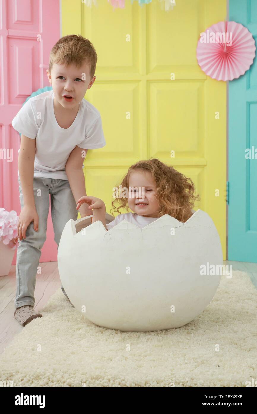 Fratello maggiore si prende cura di sua sorella piccola è seduta in un uovo bianco che si schiuse contro uno studio colorato sfondo. Concetto familiare. Foto Stock