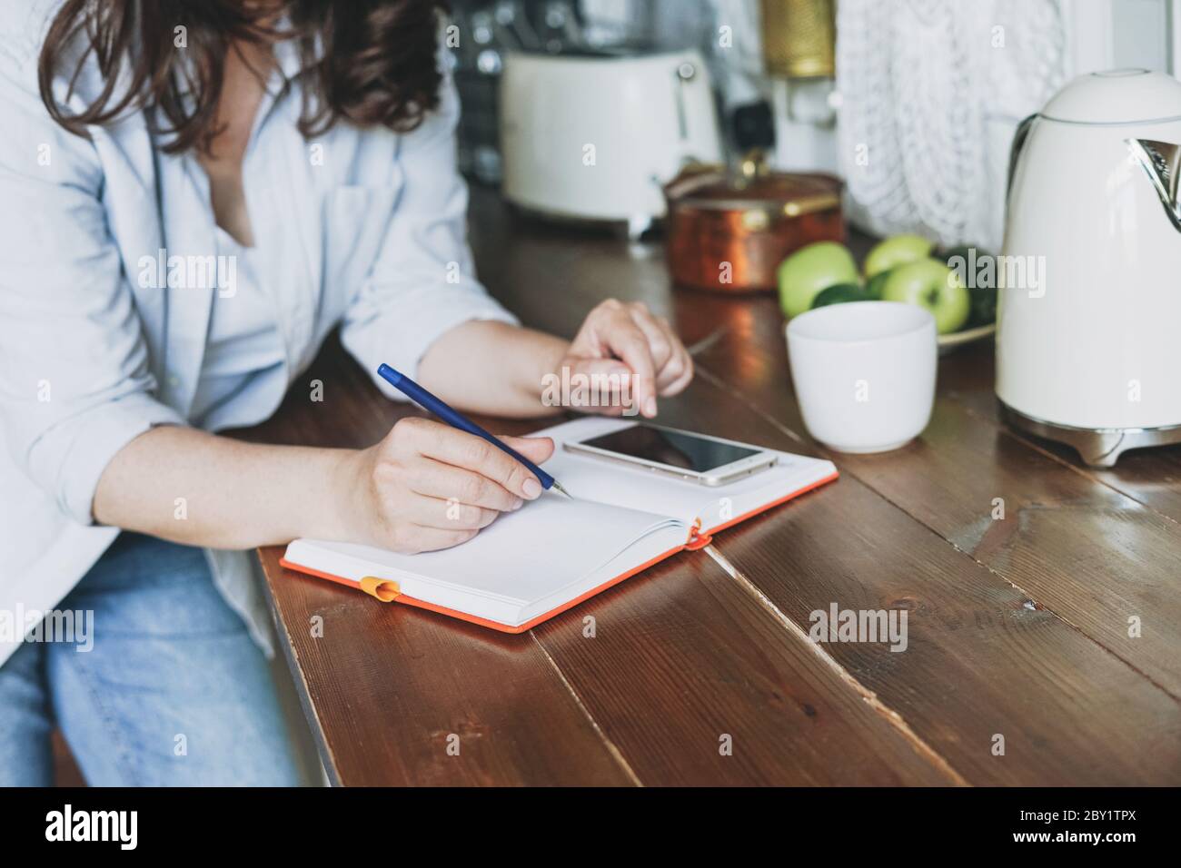 Donna brunette in informale facendo note nel libro quotidiano con e utilizzando mobile in cucina a casa Foto Stock