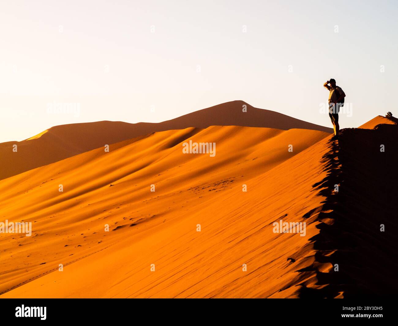 Silhouette di giovane uomo che si erge sulla cresta della duna rossa del deserto del Namib vicino a Sossusvlei al tramonto, Namib-Naukluft National Park, Namibia, Africa Foto Stock