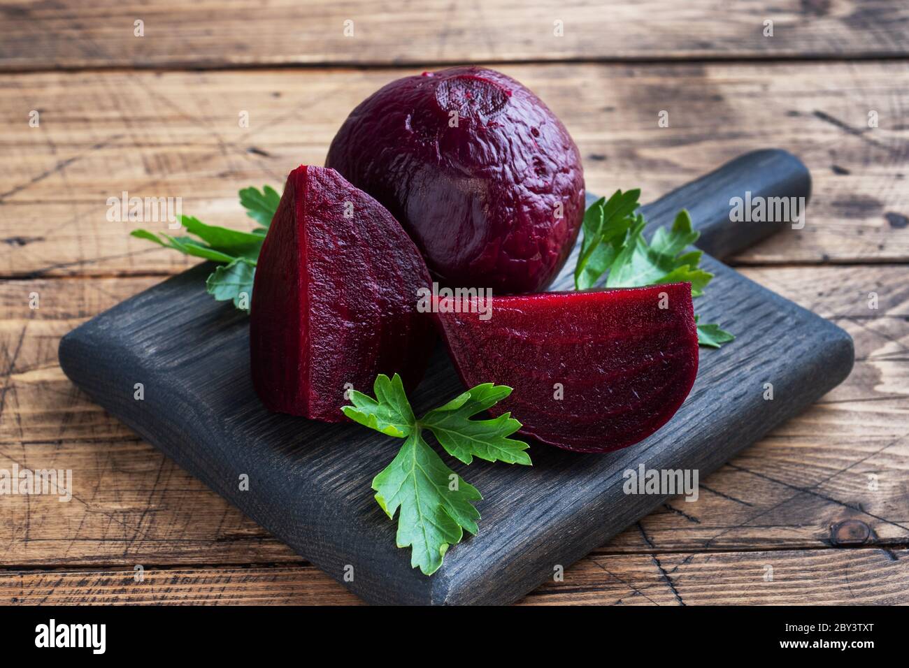 Barbabietole bollite intere e tagliate su un tagliere con foglie di prezzemolo su uno sfondo rustico di legno Copia spazio Foto Stock