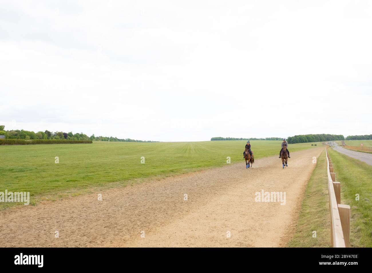 Coppia di piloti visti su cavalli da galoppo in uno spazio pubblico aperto. Vista adiacente a recinzioni e una strada deserta, che conduce dalla cima di una collina. Foto Stock