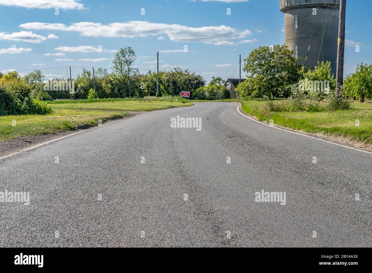 Vista a livello del suolo di una strada rurale vuota che mostra la superficie stradale testurizzata. Un cartello stradale distante si può vedere vicino ad una curva stretta, vicino ad una torre dell'acqua. Foto Stock