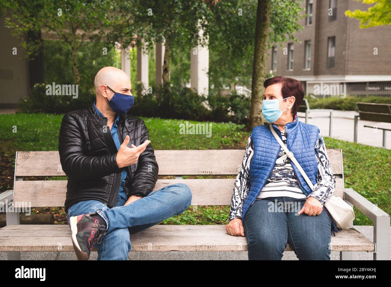 Vecchia donna con il suo figlio maturo che indossa maschera di faccia seduta su una panchina all'aperto mantenendo la distanza sociale nella nuova normalità Foto Stock