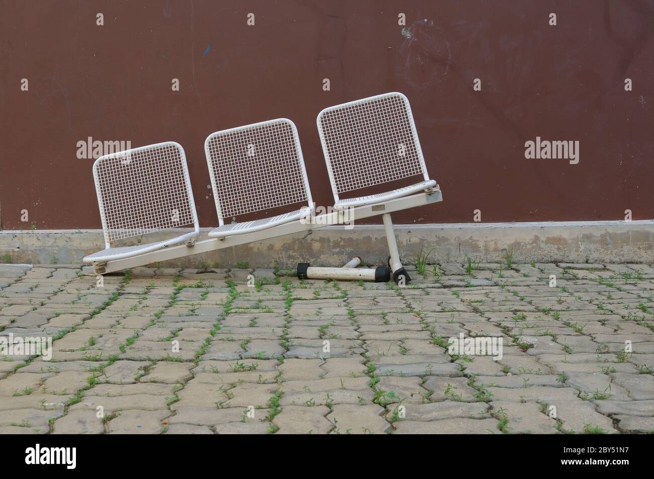 Fila di sedie in metallo bianco danneggiate con gamba rotta, sedie inclinate vengono abbandonate su pavimentazione in pietra nel parco Foto Stock