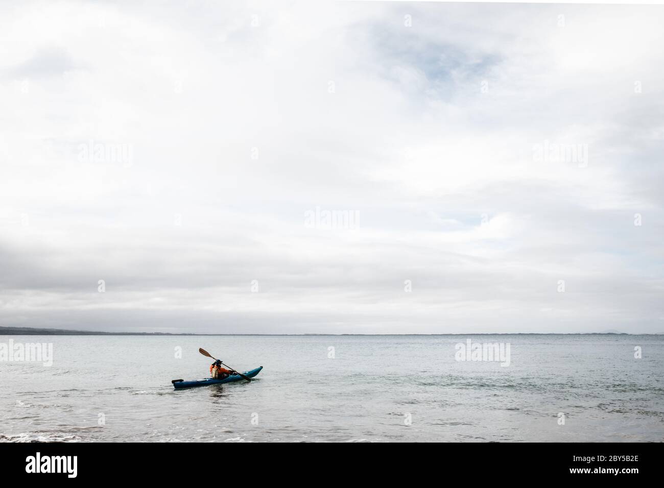 Un uomo che usa un kayak blu nell'oceano alla spiaggia di Walkerville nord in Victoria Australia, in una giornata ancora ma nuvolosa Foto Stock