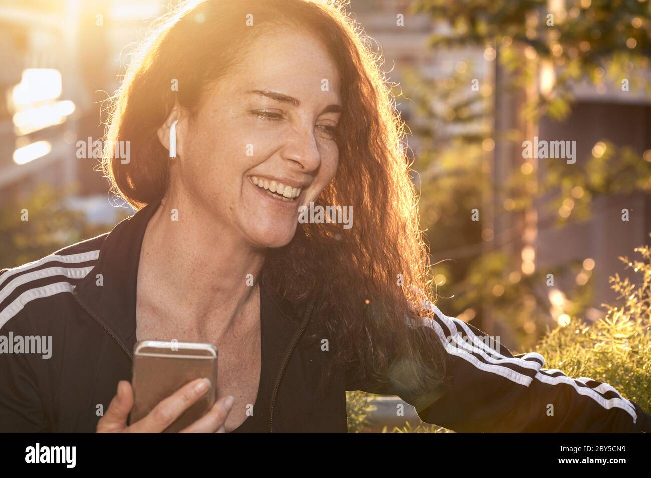 Ritratto di una donna matura che sorride parlando su un telefono cellulare utilizzando auricolari wireless all'aperto, felice, corpo in forma Foto Stock