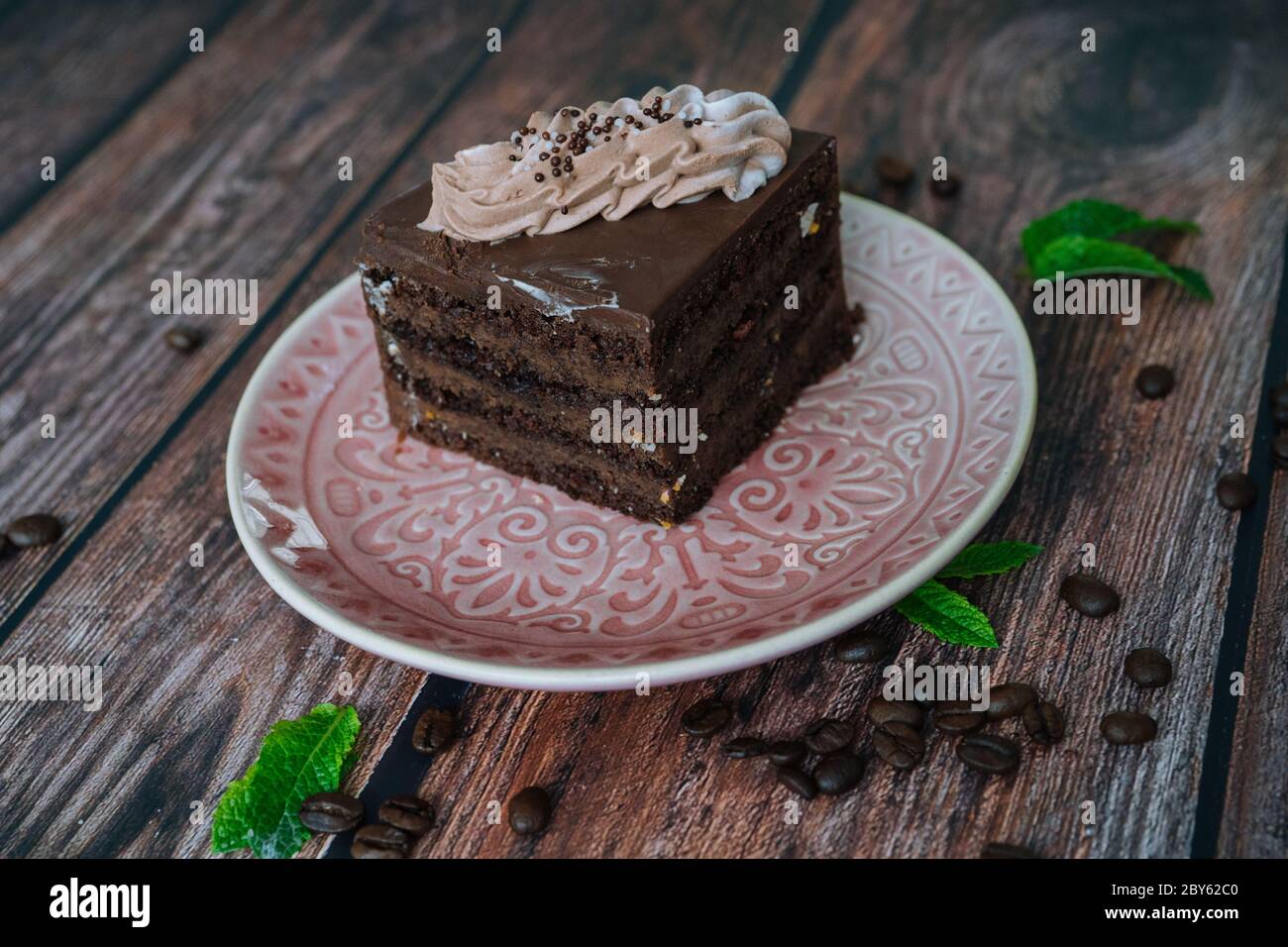 Pezzo di torta al cioccolato con crema sul piatto. Torta al cioccolato su sfondo di legno con chicchi di caffè e foglie di menta. Deliziosa fetta di torta a strati sul piatto. Cottura e decorazione torta al cioccolato Foto Stock