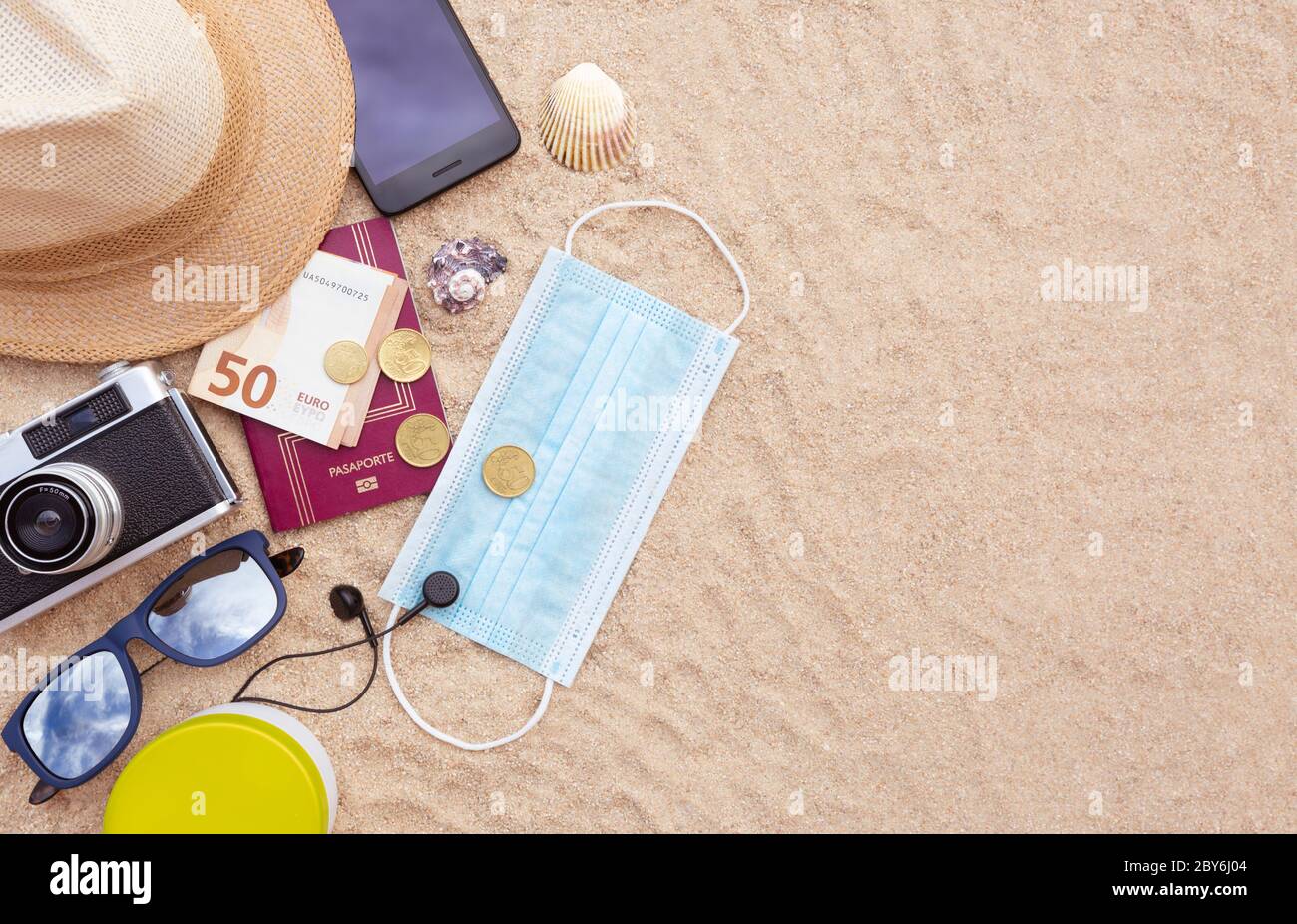 Alcuni oggetti da spiaggia, una maschera protettiva, un passaporto e alcuni contanti, uno smartphone, cappello, macchina fotografica e occhiali da sole sulla spiaggia di sabbia. Spazio per il testo. Concep Foto Stock