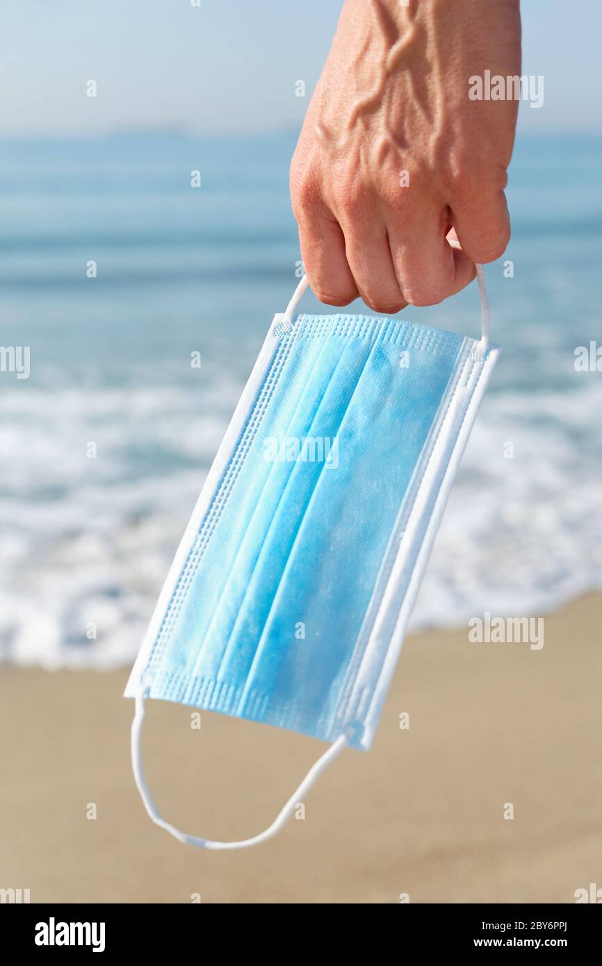 primo piano di un giovane caucasico con una maschera chirurgica blu in mano sulla spiaggia, come sta prendendo un respiro di portarla, accanto alla riva del mare Foto Stock