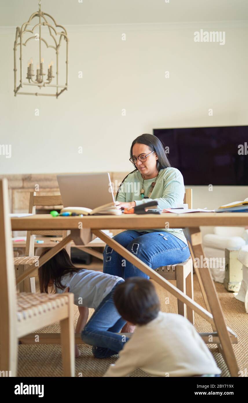 I bambini giocano sotto il tavolo mentre la madre lavora al computer portatile Foto Stock
