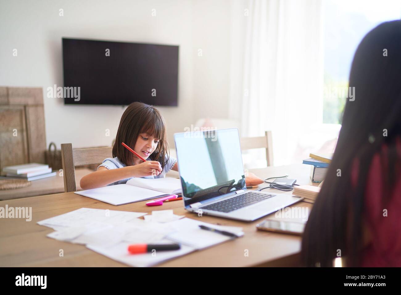 Ragazza che fa i compiti mentre la madre lavora al computer portatile sul tavolo da pranzo Foto Stock