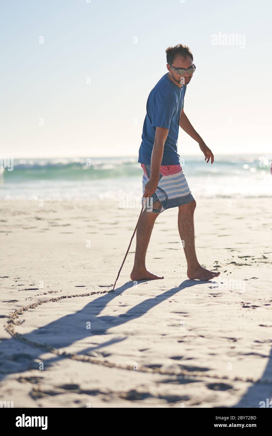 Uomo che si disegna in sabbia con bastone sulla spiaggia soleggiata Foto Stock