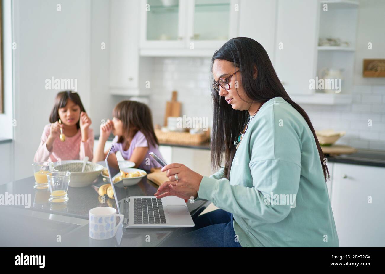 Madre che lavora al computer portatile in cucina mentre le figlie mangiano la colazione Foto Stock