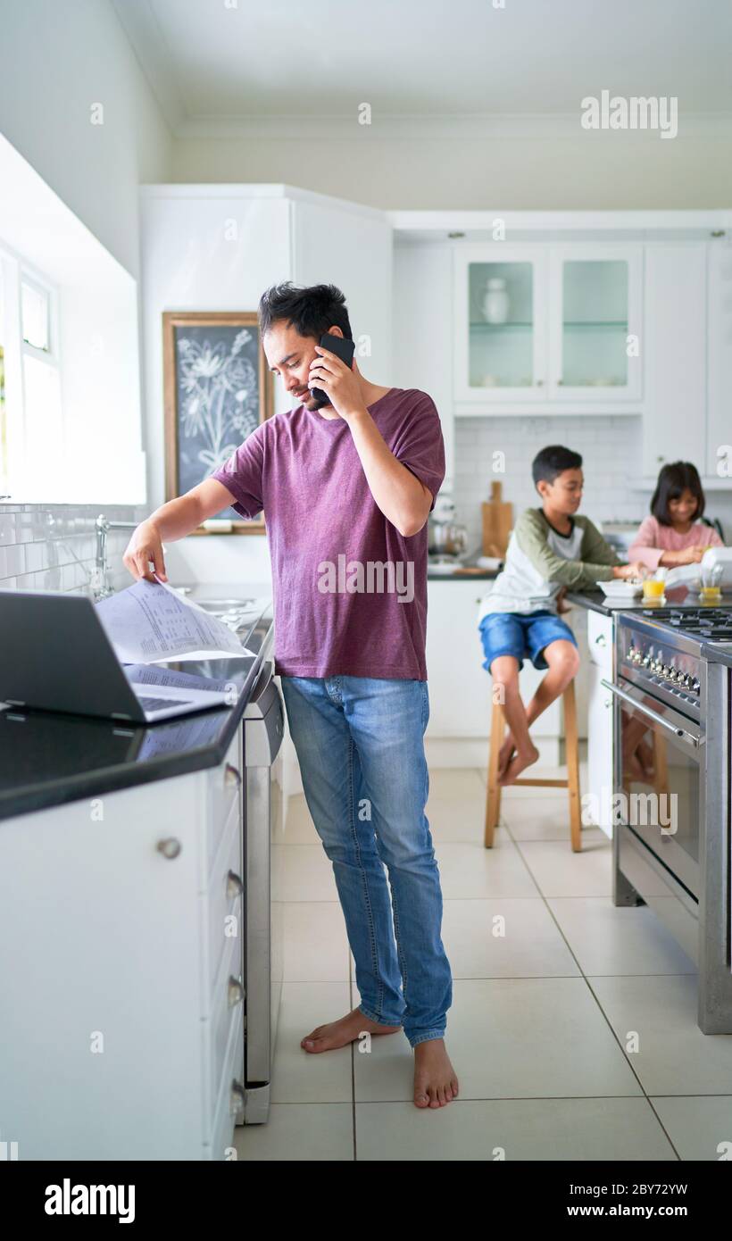 Padre che lavora al computer portatile in cucina con i bambini che mangiano Foto Stock